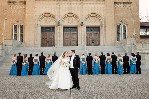 the happy couple standing in front of historic cathedral with a line of the wedding party behind them facing the cathedral- bride is wearing white ball gown style dress with full length veil and groom is wearing black and white tuxedo, and bridesmaids are wearing full length blue dresses - photo by Houston based wedding photographer Adam Nyholt